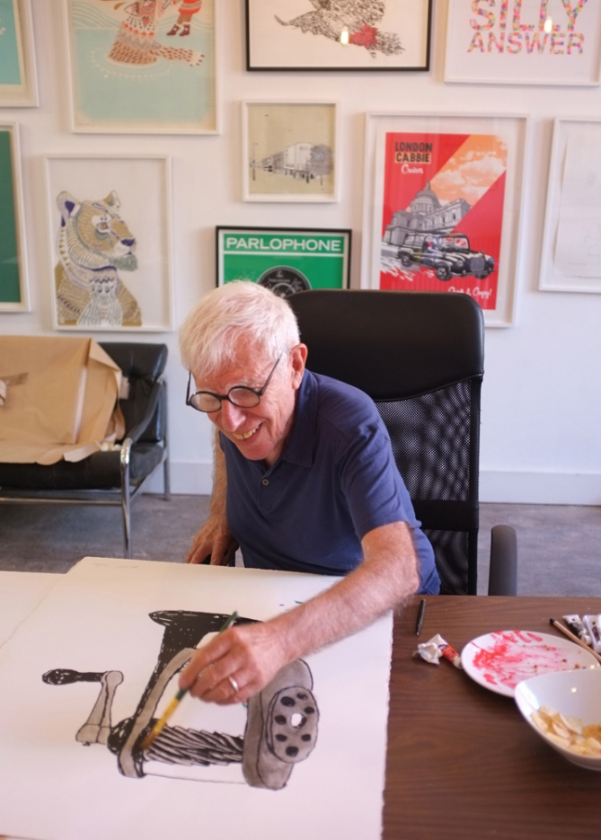 An elderly man with glasses sits at a desk, smiling whilst painting a black-and-white drawing. Framed artwork is displayed on the wall behind him.