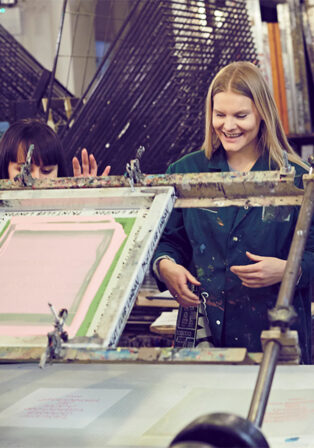 Two women smile whilst using equipment at the Beginners Screen Print Workshop (£75pp), collaborating in an art studio setting.