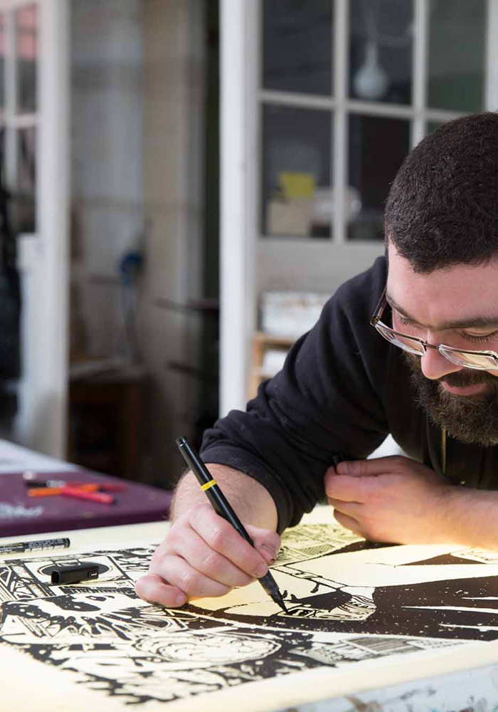 In a well-lit studio, a person with glasses uses a pen to create artwork during the Beginners’ Screen Print Workshop (£75pp).
