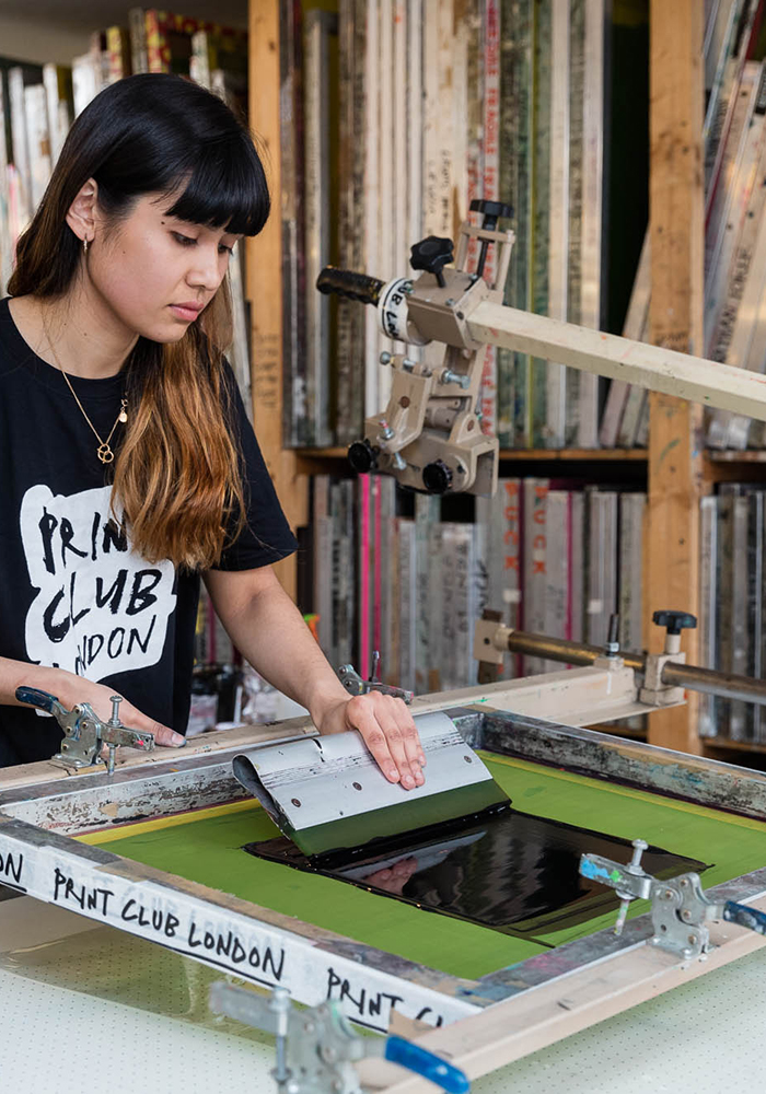 A woman takes part in the Beginners’ Workshop (£75pp), using a screen-printing press to apply black ink onto fabric in a studio, with shelves of frames and equipment visible behind her.