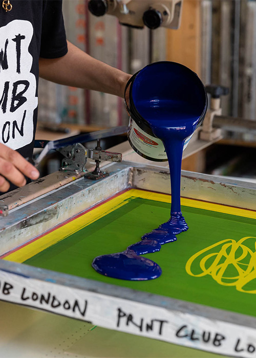 A person prepares to create a vibrant print during the Deluxe Digital Workshop (£160pp) by pouring blue ink onto a screen printing frame in a studio.
