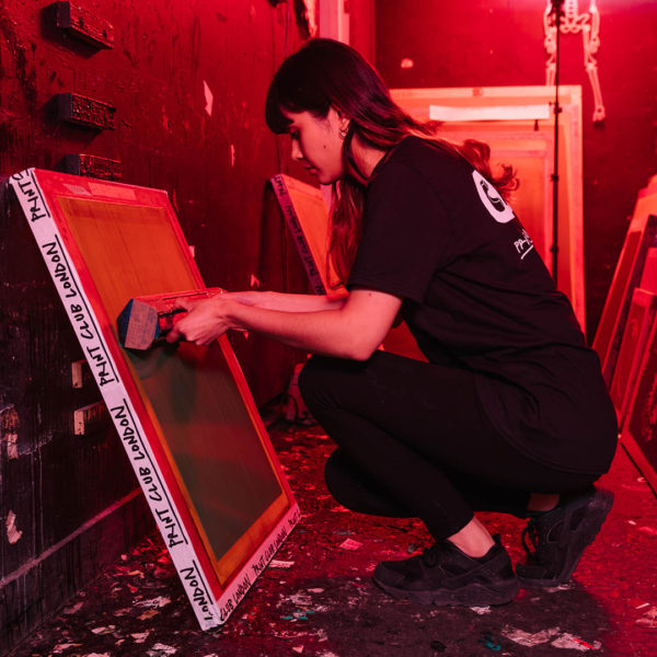 In a dimly lit studio, an artist kneels with focused intent, meticulously preparing the screen exposure for printing. The room is an art haven, filled with various screens and scattered paint splatters, each marking moments of inspiration. The ambiance is one of concentrated creativity—a sanctuary where every piece is uniquely crafted by the artist's skilled hands and imaginative spirit.