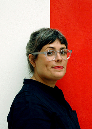 A person with grey-streaked hair, clear glasses, and a dark top stands before a wall divided into white and red sections, showcasing original artwork, whilst looking at the camera.