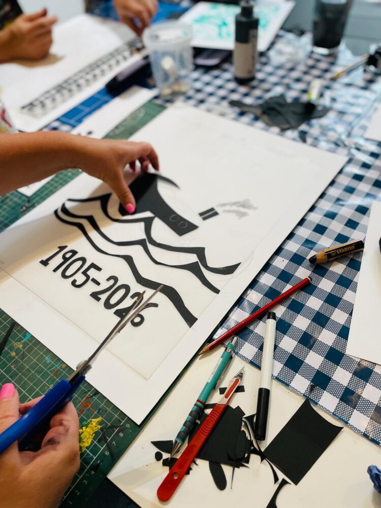 At a craft table covered with art supplies and patterned cloth, a person carefully cuts out a black paper stencil of a boat and the years 1905–2016, preparing materials for the 2 Day Screen Printing Workshop (Design Handmade) in London (£160pp).