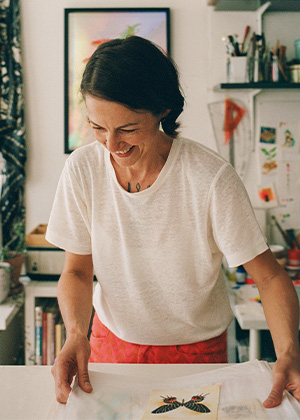 A woman in a white shirt smiles while working on an art project in a room filled with art supplies and colorful artwork.