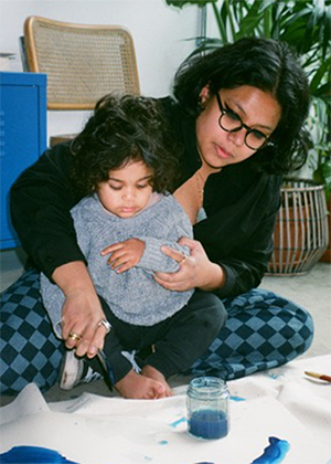 An adult helps a young child paint on paper with blue paint while sitting on the floor indoors.