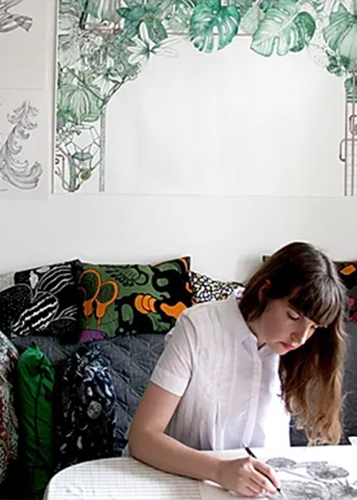 A woman sits at a table, drawing on paper, with patterned pillows and botanical-themed artwork on the wall behind her.