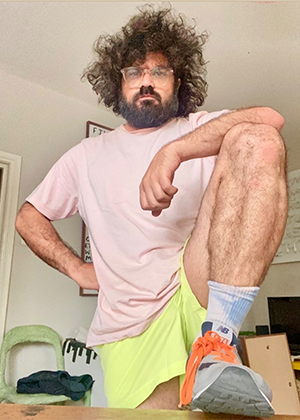 A person with curly hair and glasses poses with one foot on a surface, wearing a light pink t-shirt, yellow shorts, blue socks, and gray sneakers with orange laces indoors.