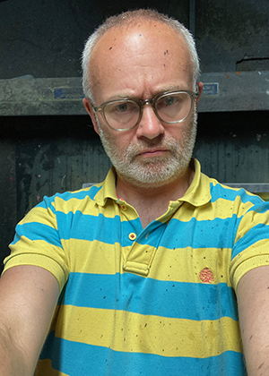 A man with glasses and a receding hairline wearing a yellow and blue striped polo shirt stands indoors, looking directly at the camera with a serious expression.