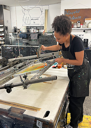 A woman wearing an apron uses a squeegee to print onto a bicycle frame in an art studio with supplies and artwork visible in the background.