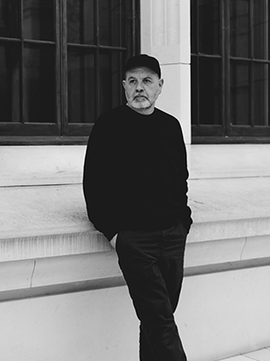 In a scene that echoes the enduring aesthetic of Anthony Burrill, a man dons a black sweater and cap as he leans nonchalantly against a white stone wall adorned with expansive windows. The monochrome composition captures a sense of timelessness and introspection, transforming the moment into a work of art.