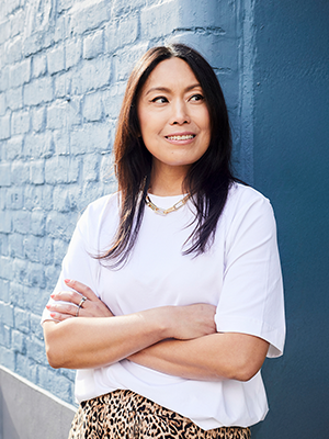 In this artwork, Cassandra Yap is depicted as a portrait of modern elegance and poise. She stands confidently against a textured blue brick wall, the rich tones of which complement her serene presence. Clad in a crisp white shirt paired with an eye-catching leopard print skirt, her outfit strikingly contrasts yet harmoniously blends with the backdrop. Her long, dark hair cascades gracefully around her shoulders, framing a gentle smile that conveys warmth and authenticity. With arms crossed casually across her chest, she exudes an aura of self-assurance and ease. The soft lighting bathes the scene in a gentle glow, enhancing the overall relaxed and inviting atmosphere of this contemporary representation.