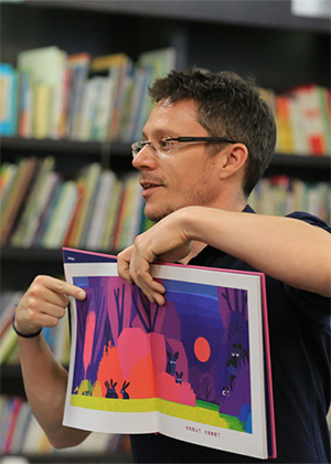 A person holding an open children's book and pointing to an illustrated page, with bookshelves in the background.