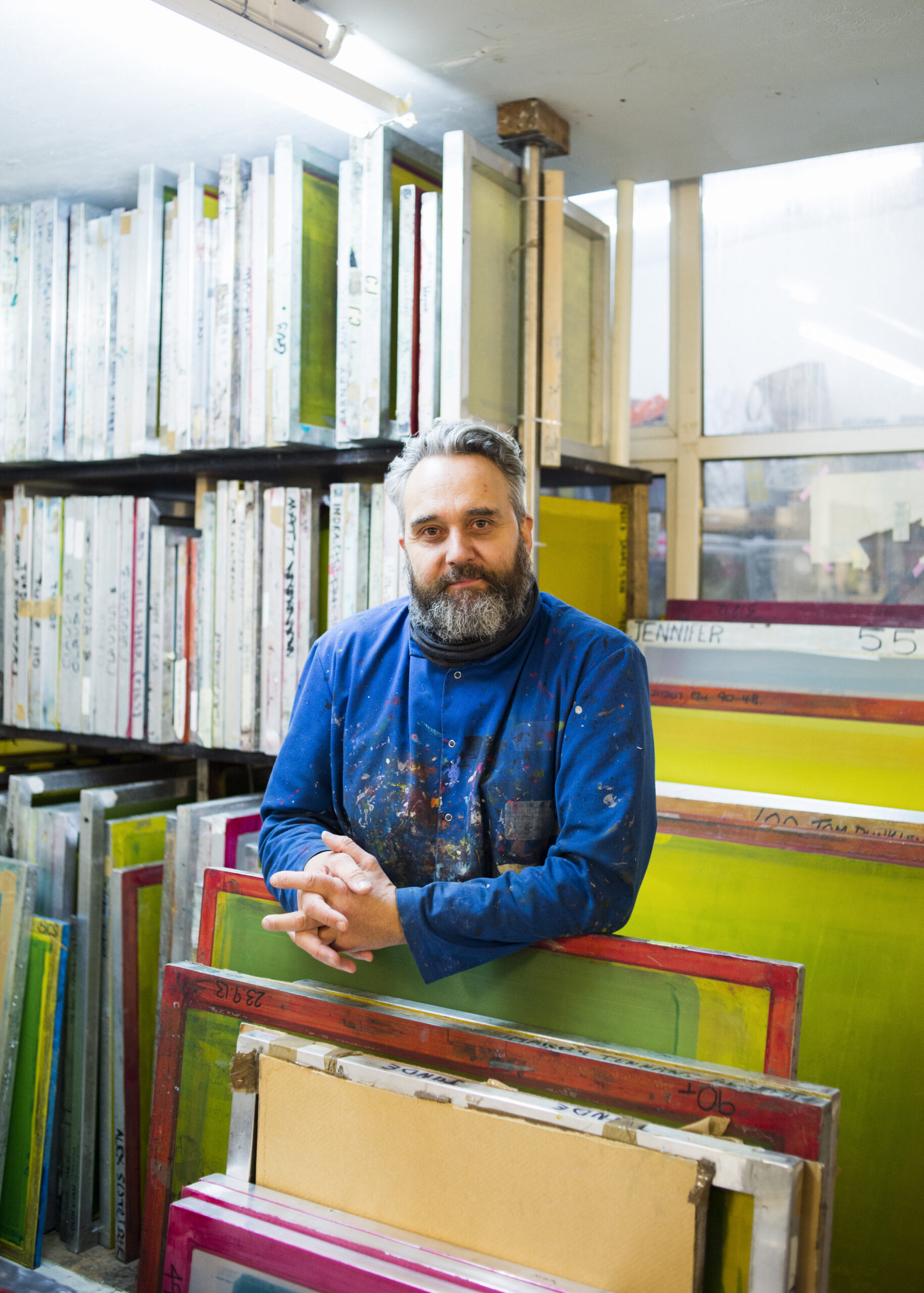 Dave Buonaguidi, a bearded man, stands wearing a paint-splattered blue shirt, surrounded by colourful screen printing frames in his vibrant workshop.