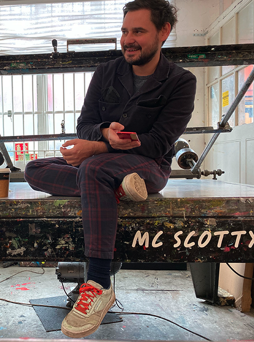A man with dark hair, wearing a navy jacket and plaid pants, sits on a paint-splattered platform holding a phone. The name "MC SCOTTY" is written on the surface, while David Newton’s influence can be seen in the creative setting.