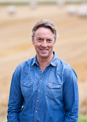 A man wearing a blue denim shirt stands outdoors in a field with blurred hay bales and golden grass in the background.