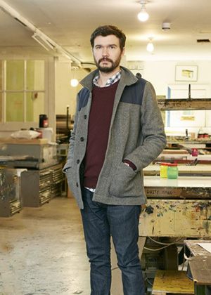 A man with a beard stands indoors wearing a grey coat, maroon jumper, checked shirt, and jeans in an art studio setting with tables and art supplies in the background.