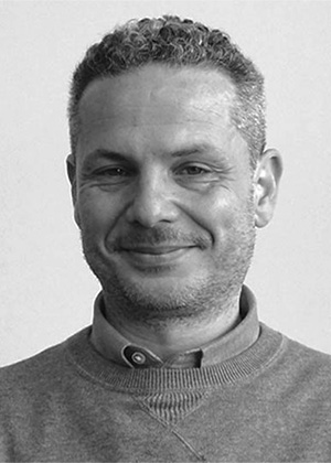 A man with short curly hair and a trimmed beard is wearing a buttoned-up shirt and a jumper, smiling at the camera against a plain background.