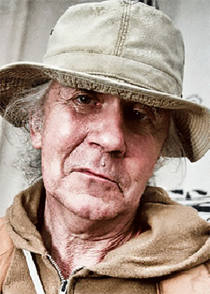 Older man with grey hair wearing a beige bucket hat and tan jacket looks at the camera with a neutral expression.