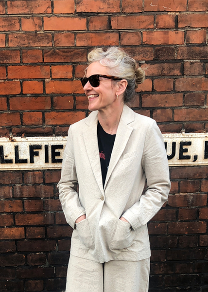 A person with short gray hair wearing sunglasses and a light-colored suit stands smiling in front of a brick wall and a partial street sign.