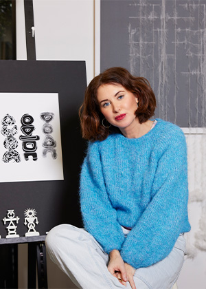 A woman with short brown hair in a blue sweater sits by artwork on an easel featuring abstract black and white designs.