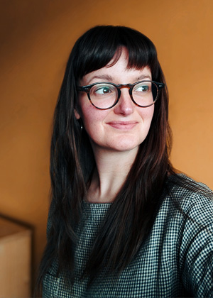 Woman with long brown hair and glasses, wearing a checked top, looks to the side and smiles slightly. The background is plain and orange-toned.