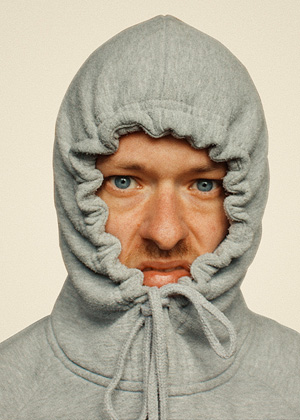 A person with blue eyes wears a grey hoodie pulled tightly around their face, with only the eyes, nose, and mouth visible.