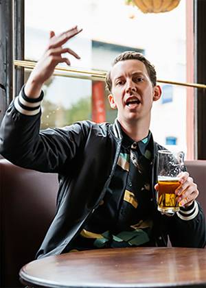 A man sitting at a table in a pub, holding a glass of beer in one hand and gesturing with the other.