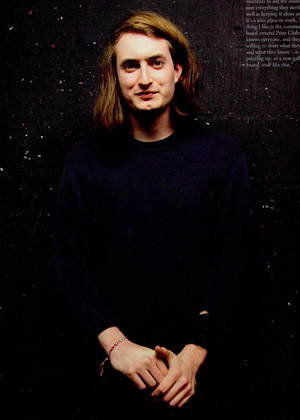 A person with long brown hair, wearing a dark jumper, stands against a dark speckled background, looking at the camera with hands clasped.