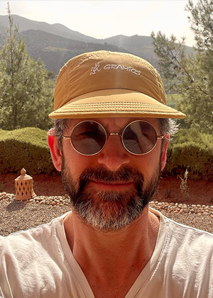 A man with a beard wearing round sunglasses and a tan cap stands outdoors with trees and mountains in the background.