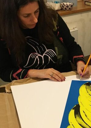 A woman sits at a table, drawing on a sheet of white paper that partially covers a blue and yellow artwork. She is holding a pencil.