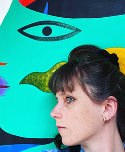 A woman with brown hair and earrings is in profile in front of a colorful abstract painting featuring a stylized face.