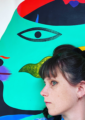 A woman with brown hair faces sideways in front of a colorful abstract painting by Margaux Carpentier that features a large stylized face.