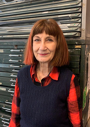 Nurit Manor, with shoulder-length brown hair and a black sweater over a red plaid shirt, stands in front of a stack of steel drawers, smiling warmly at the camera.