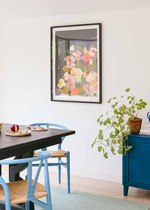 In this composition, the dining area is transformed into a harmonious canvas of contrasts and complements. At the center stands a dark wooden table, its rich tones providing depth and grounding to the scene. Encircling it are chairs adorned in vibrant blue, which echo both serenity and innovation. The wall behind serves as a gallery space for a colorful abstract painting by Print Club London, infusing an element of dynamic creativity with its lively hues.  A solitary potted plant rests atop a blue cabinet, introducing a touch of natural elegance and freshness to the tableau. This botanical accent not only enhances the aesthetic but also bridges indoor comfort with organic life. Natural light pours into this serene setting, illuminating each element to create an inviting symphony of color, texture, and form where modern art meets everyday functionality.