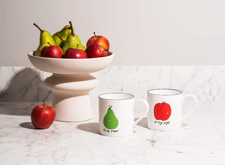 In the style of a Print Club London masterpiece, a stone table is artfully arranged with a bowl overflowing with vibrant red apples and lush green pears. Accompanying this colorful bounty are two white mugs: one adorned with an illustration of a green pear and the words "Wise Pear," while the other displays a striking red apple and the phrase "of my eye." Completing this tranquil composition, a solitary red apple sits contemplatively on the table surface—a harmonious arrangement that captures warmth and familiarity in every detail.