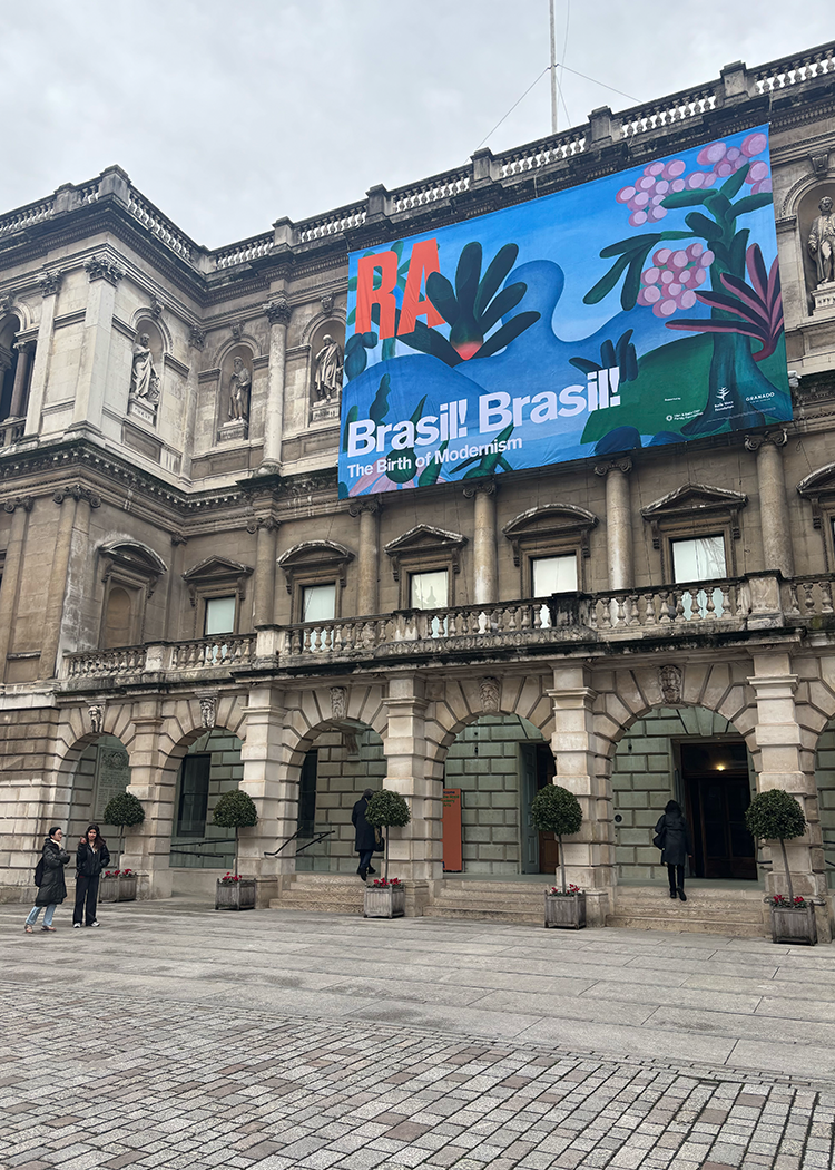 The historic building's exterior, a masterpiece of classical architecture, serves as the canvas for the exhibition "RA Brasil: The Birth of Modernism" at the Royal Academy. A lively banner adorned with colorful plant motifs welcomes visitors, while clusters of people gather near the grand entrance. Majestic potted trees encircle the courtyard, adding a touch of natural grace to this artistic setting.