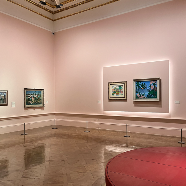 In this serene art gallery, light pink walls evoke the essence of Modernism, providing a gentle backdrop for four framed paintings. The space boasts elegant wooden floors and intricate decorative molding. A striking red circular bench occupies the foreground, inviting visitors to pause and reflect. Subtle lighting illuminates the artworks, capturing the vibrant creativity and spirit of Brazil.