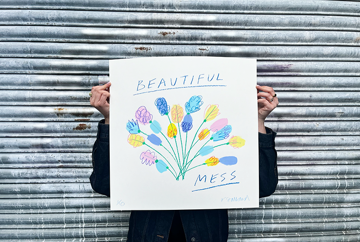 An artistic portrait captures an individual posed against a textured, corrugated metal background, holding a vibrant piece from Print Club London. This striking artwork showcases a symphony of colorful abstract flowers with the words "BEAUTIFUL MESS" gracefully inscribed both above and below. The person's dark jacket provides a stark contrast to the lively hues of the sign, highlighting the chaotic beauty embodied in the message and design.