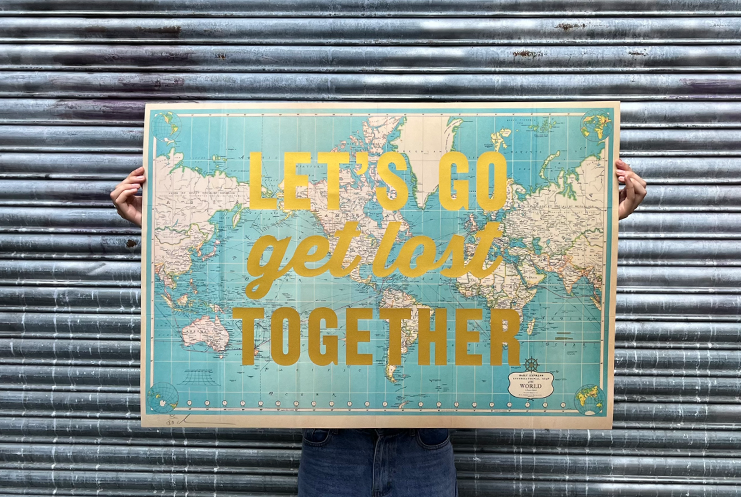 A person stands before a metal wall. They hold a world map. Bold, golden letters shout: "LET'S GO get lost TOGETHER." This striking piece comes from Print Club London.