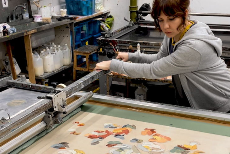 A person in a gray hoodie runs a screen printing machine. The workshop feels like Print Club London. A canvas with colorful abstract patterns lies on the table. Shelves in the background hold supplies. Bottles and folded fabrics fill the shelves.