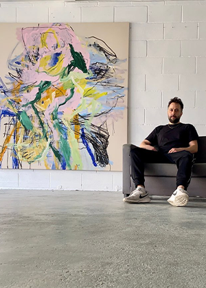 A man in casual clothing sits on a gray couch next to a large, colorful abstract painting by Ben Wakeling in a minimalist room with a white brick wall.
