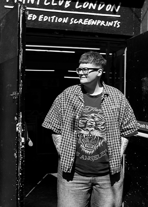 Caitlin Flood-Molyneux, wearing glasses, a plaid shirt, and a graphic t-shirt, stands in the doorway of a building labeled "Print Club London Limited Edition Screenprints.