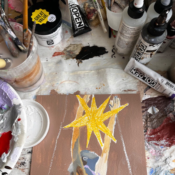 A painting in progress on a cluttered art table, surrounded by brushes, paint tubes, mixing containers, and art supplies. The painting features a yellow star and raised arms.