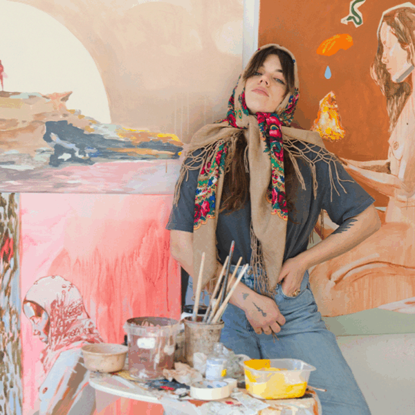 A woman in a headscarf leans against a wall with colorful paintings behind her and art supplies on a table in front.