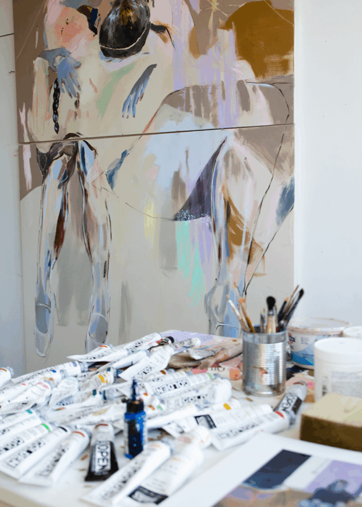 A table with paint tubes, brushes, and art supplies sits in front of an abstract painting of two human figures on a studio wall.