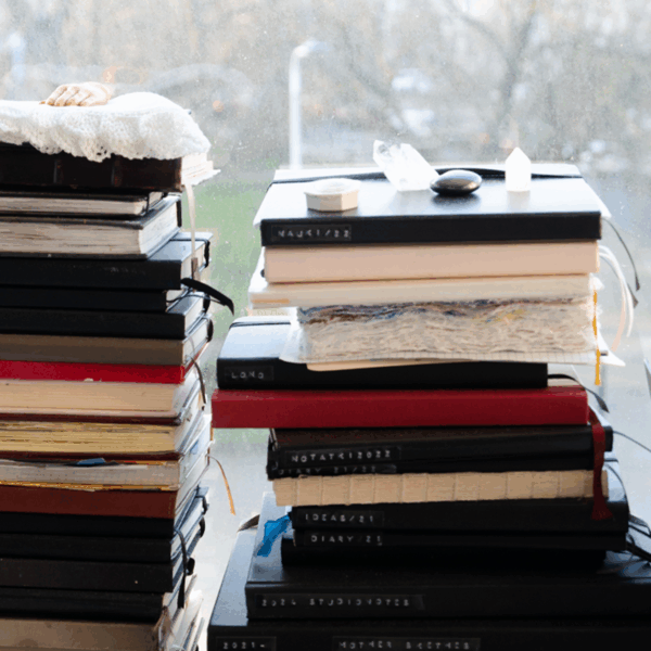 Two tall stacks of assorted notebooks and journals sit on a windowsill, with small objects and a doll's hand on top of the piles. Trees and a street are visible outside.