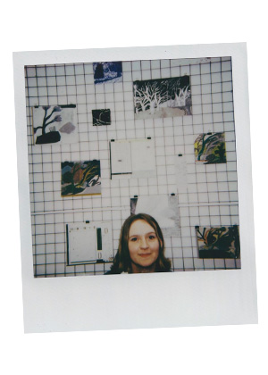 A woman, likely Chantelle Wroblewska-Fairless, stands in front of a grid wall displaying various small artworks and papers, with only her head visible in the lower center of the image.