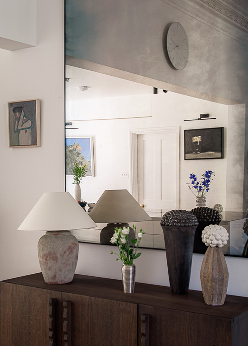 A large mirror reflects a white wall adorned with Print Club London artwork and a clock. On the dark cabinet below, four vases with flowers and a lamp complete the stylish scene.