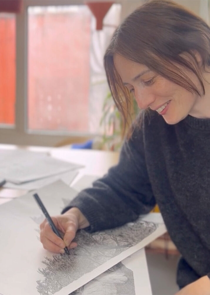 A woman sits at a table, smiling as she draws with a pen on detailed paper artwork in a well-lit room.